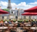 Café parisiense com mesas e cadeiras de vime sob guarda-sóis vermelhos e Torre Eiffel ao fundo.