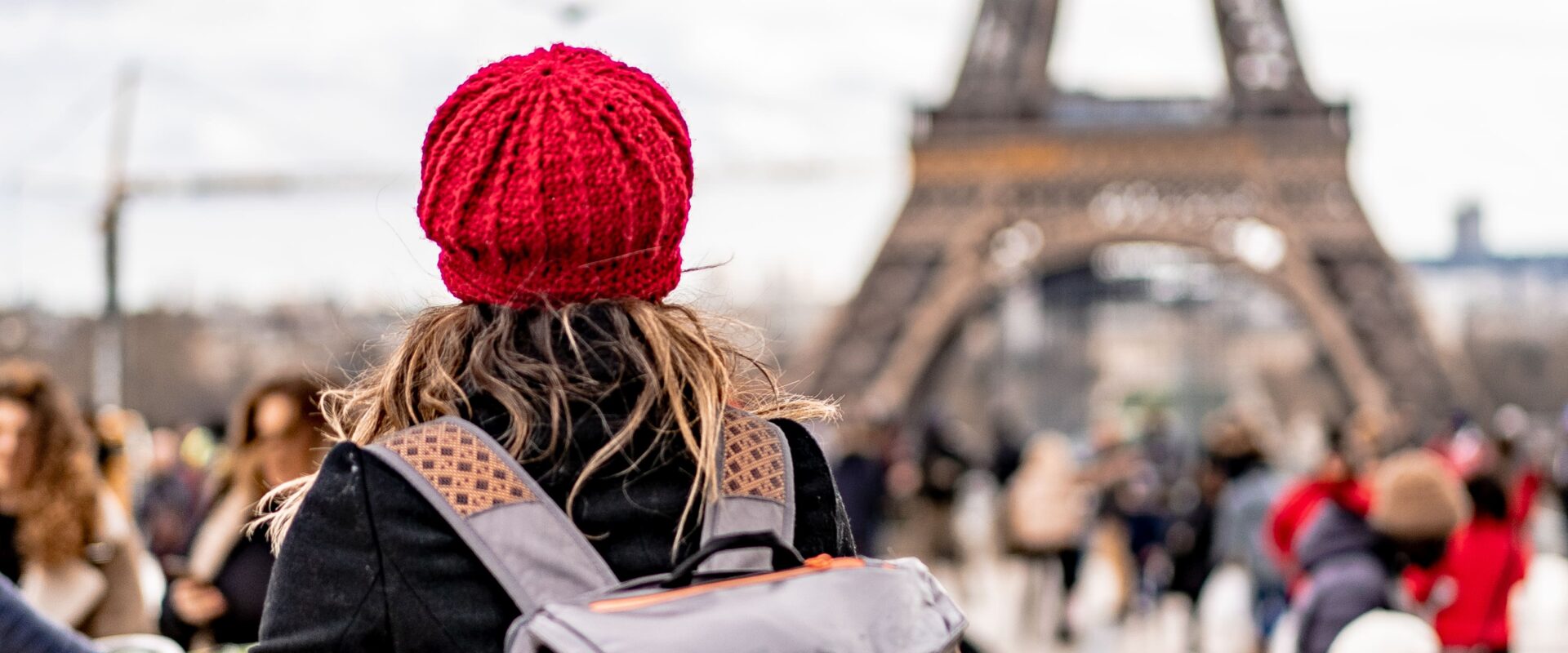 Uma pessoa está em frente à Torre Eiffel, em Paris, observando o monumento de costas para a câmera. Ela usa um casaco escuro e carrega uma mochila cinza nas costas, além de levar uma bolsa ao lado do corpo. A pessoa está em uma área movimentada, com vários turistas ao redor. A Torre Eiffel aparece ao fundo, ocupando grande parte da imagem e se destacando contra um céu claro. A cena representa uma viagem turística a Paris.