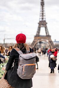 Uma pessoa está em frente à Torre Eiffel, em Paris, observando o monumento de costas para a câmera. Ela usa um casaco escuro e carrega uma mochila cinza nas costas, além de levar uma bolsa ao lado do corpo. A pessoa está em uma área movimentada, com vários turistas ao redor. A Torre Eiffel aparece ao fundo, ocupando grande parte da imagem e se destacando contra um céu claro. A cena representa uma viagem turística a Paris.